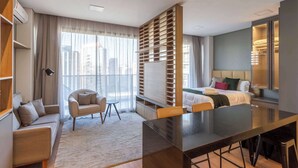 Una habitación de hotel moderna con sofá, mesa de comedor, cama, armario y un amplio ventanal con vista a la ciudad.
