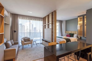 Ein modernes Hotelzimmer mit einer Couch, einem Esstisch, einem Bett, einem Kleiderschrank und einem großen Fenster mit Blick auf die Stadt.