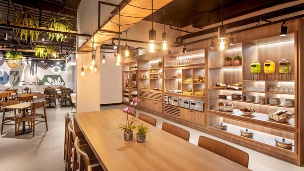 Un ristorante con tavoli e sedie in legno, lampade a sospensione e scaffali esposti con cibo e bevande.