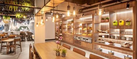 Un restaurante con mesas y sillas de madera, lámparas colgantes y estantes con productos alimenticios y bebidas.