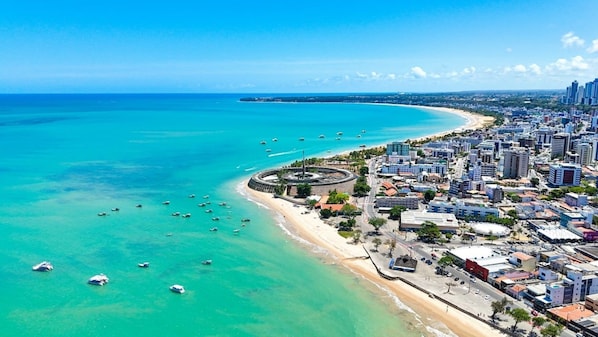 An aerial view of a coastal city with a prominent circular structure near the beach, dense urban development, and clear turquoise waters.