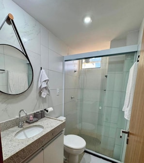 A modern bathroom with a glass shower enclosure, a sink with a mirror, and a toilet. - Flat Santorini com Piscina - João Pessoa (João Pessoa)