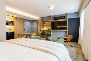A modern hotel room with a bed, a desk, a chair, a TV, and a shelf with books and a lamp. - #EB904 | Cyclinn Brooklin Santo Amaro (Brooklin)
