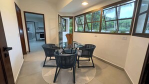 Un coin repas avec une table en verre et des chaises en osier, un vase posé sur la table et de grandes fenêtres donnant sur un paysage verdoyant.