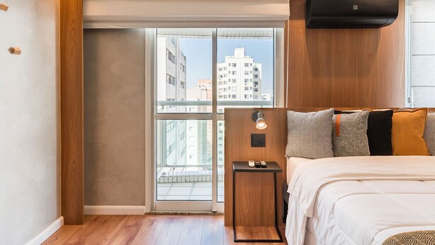 Ein modernes Hotelzimmer mit großem Fenster, einem Bett, einem Nachttisch mit Lampe und einer Wand-Klimaanlage.