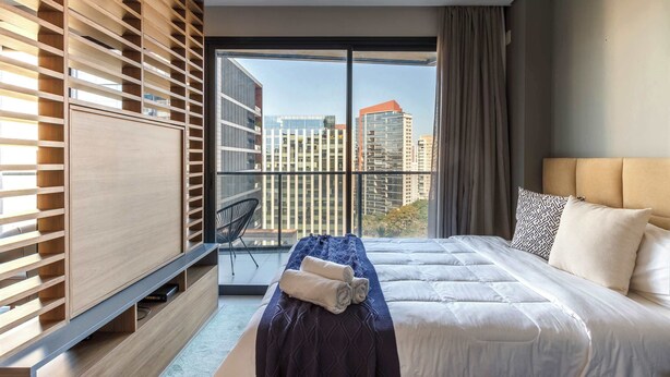 A modern hotel room with a large window, a bed with white bedding, a wooden shelf unit, and a massage table. - #VN1711 | Cyclinn Vila Olímpia (Vila Olímpia)