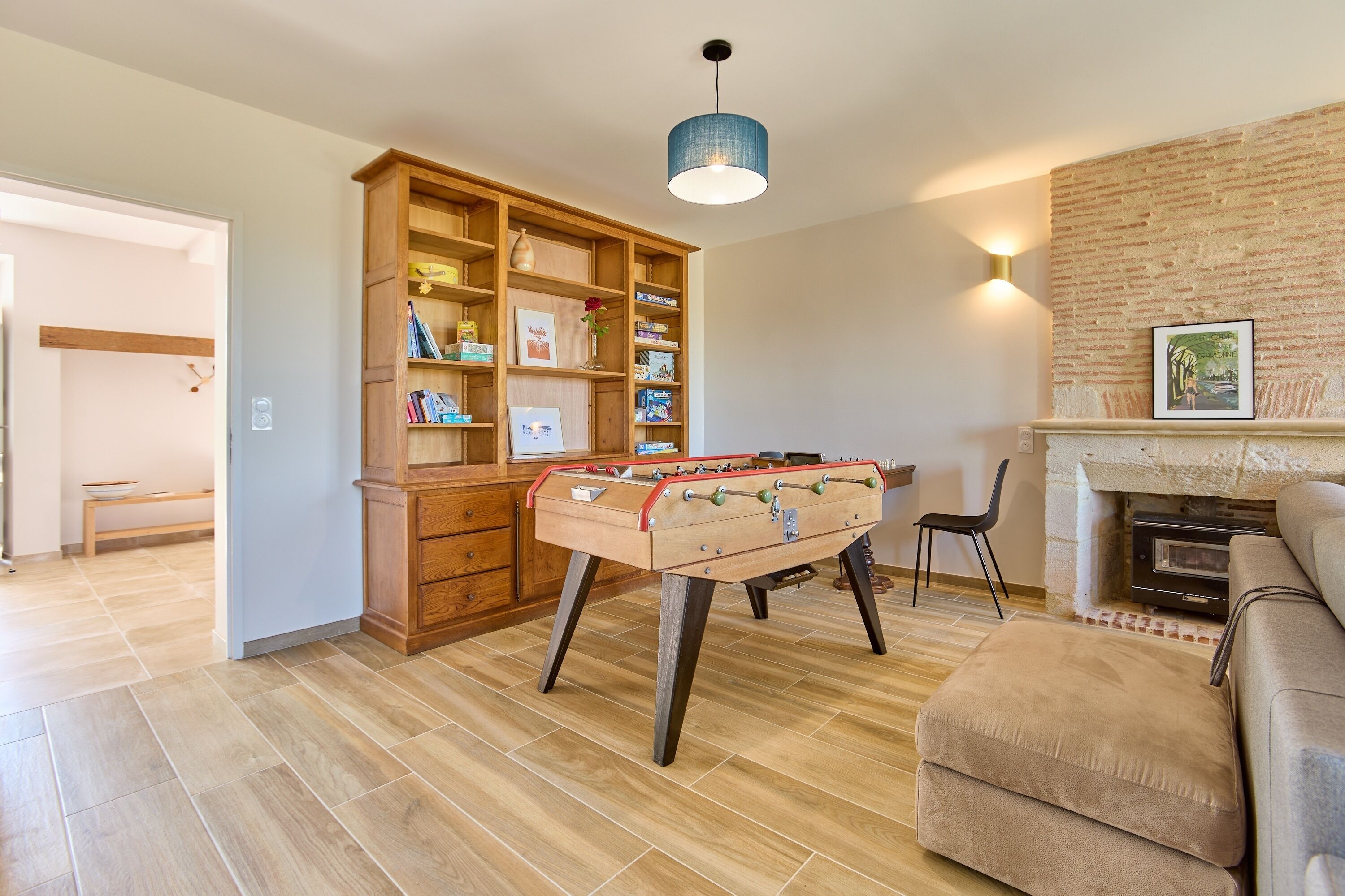 Ein gemütliches Wohnzimmer mit Kicker, einer Holzbücherreol, einem Kamin und einer modernen Pendelleuchte.