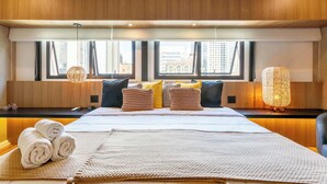 Ein Hotelzimmer mit einem großen Bett, mehreren Kissen, einem Nachttisch, einer Lampe und Blick auf die Stadt durch große Fenster.
