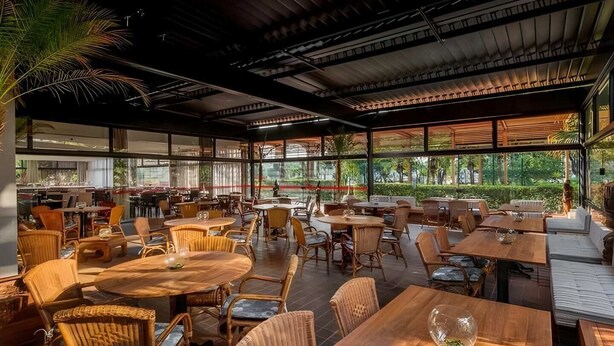 An indoor/outdoor dining area with multiple tables and chairs, large windows, and a view of greenery outside. - #TB413 | Cyclinn Berrini Brooklin (Berrini)