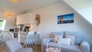 Ett modernt vardagsrum med en grå soffa, ett litet bord med koppar, en vit stol och en köksdel med vita skåp och ett matbord.