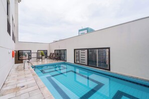 Una piscina en la azotea con suelo de baldosas, sillones de descanso y vistas a edificios.