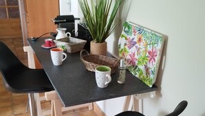 Angolo cottura con piano di lavoro nero, due sedie nere, una pianta in vaso, un quadro floreale e diversi oggetti da cucina.