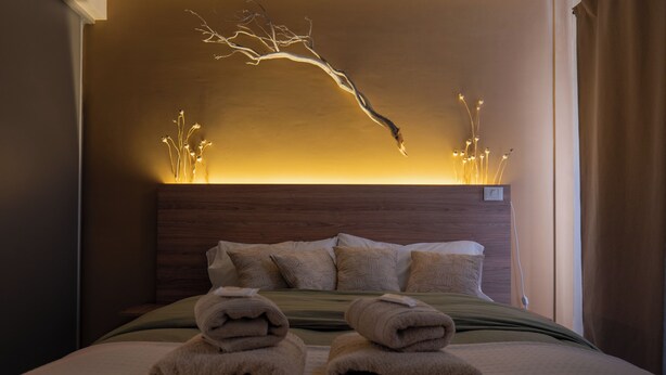 Ein ordentlich bezogenes Bett mit gefalteten Handtüchern, einem Holz-Kopfteil und einer Wandleuchte.