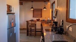 Eine Küche mit Kühlschrank, Holzkstühlen, einem Esstisch, Pendelleuchten, einer Kaffeemaschine und einem Fenster.