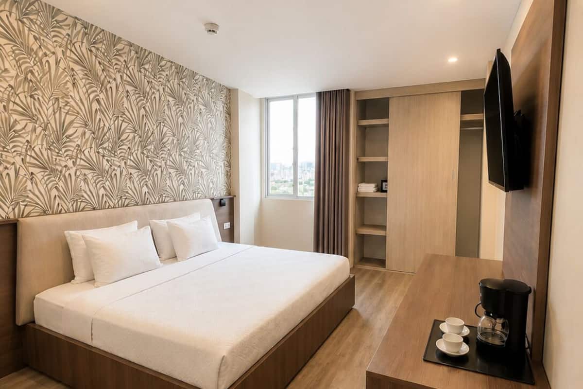 A modern hotel room with a large bed, a desk with a coffee maker, a TV, and a wardrobe.