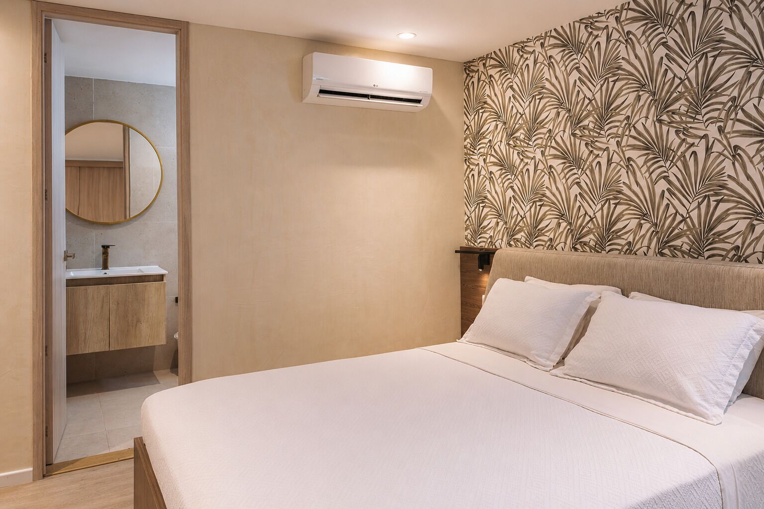 Un dormitorio con cama, aire acondicionado instalado en la pared, baño con espejo y lavamanos, y papel tapiz estampado.