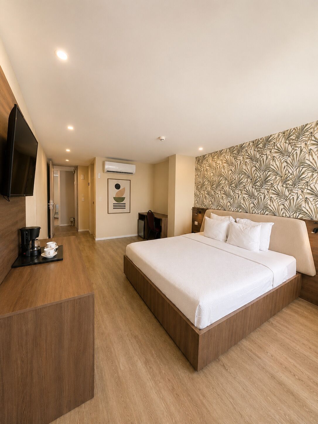A hotel room with a large bed, a desk with a coffee maker, a TV, and an air conditioning unit.