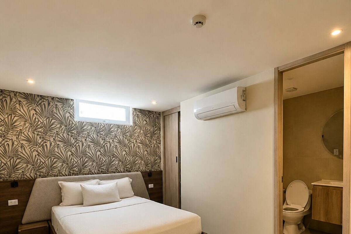 A bedroom with a bed, a window, an air conditioning unit, and a bathroom visible through an open door.