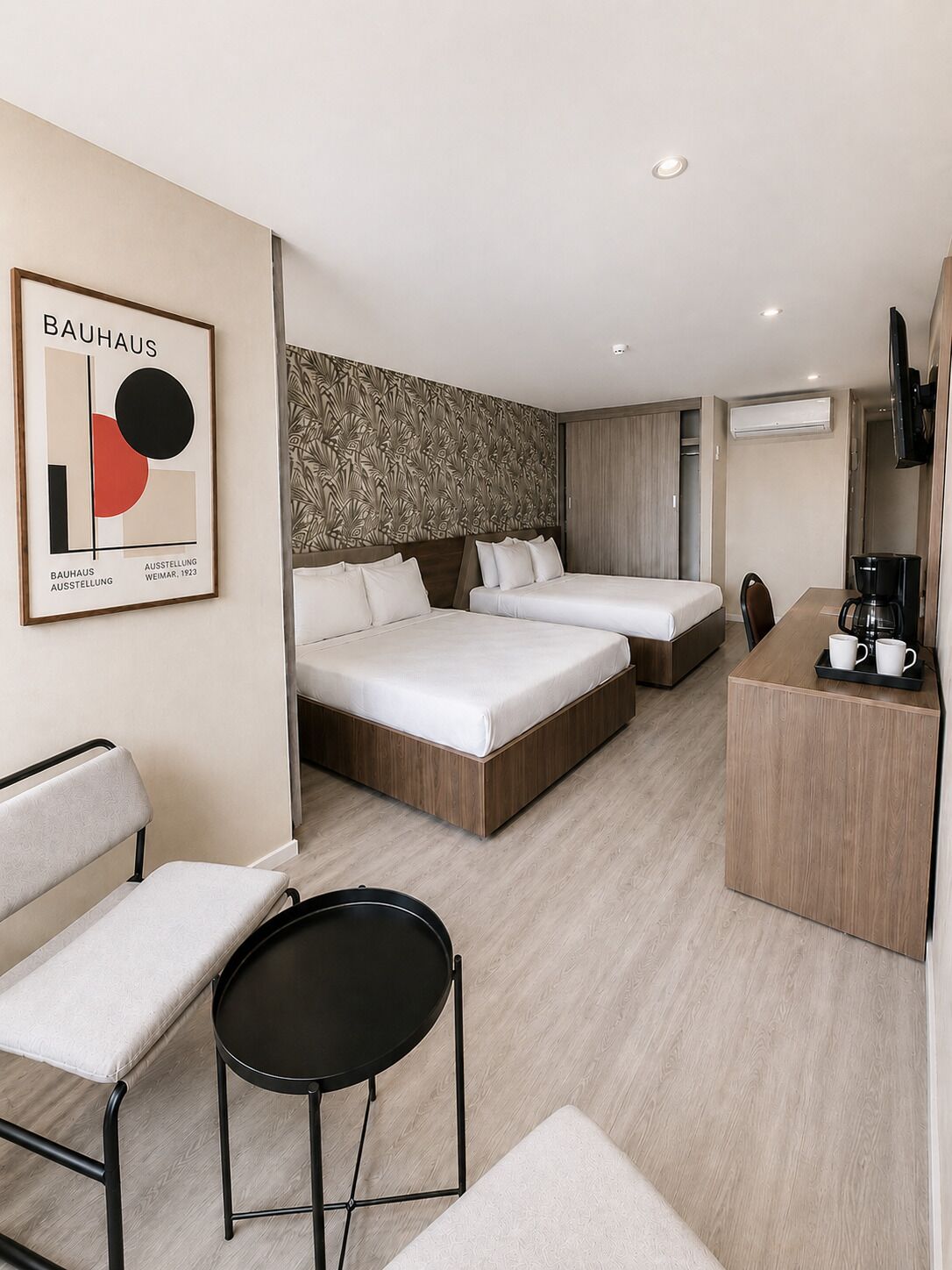 A modern hotel room with a large bed, a smaller bed, a desk with a coffee maker, and a TV mounted on the wall.