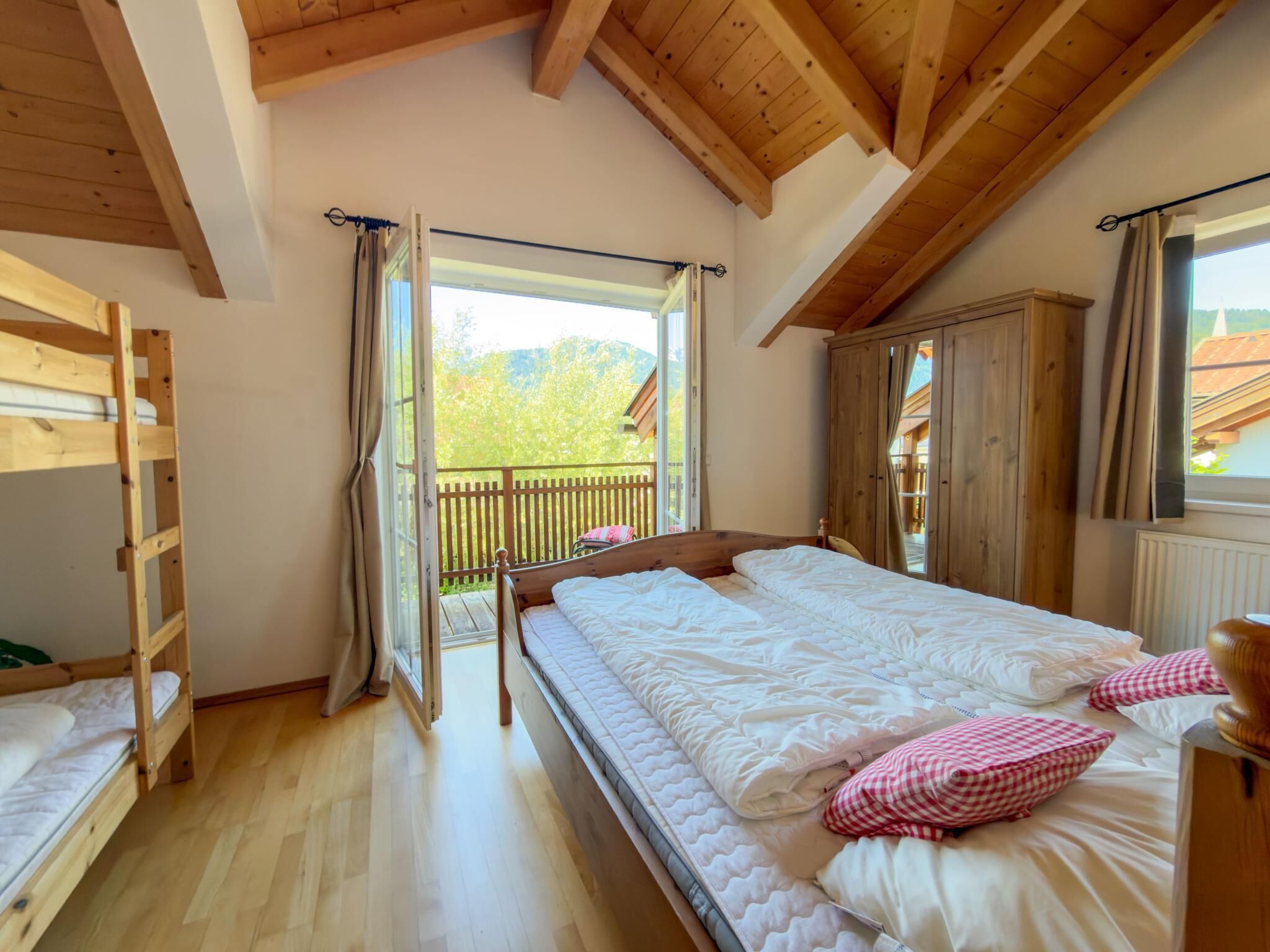 Ein Schlafzimmer mit Holzboden, einem Bett mit weißer Bettwäsche, einem Holzschrank und einem Balkon mit Blick auf Grünflächen.