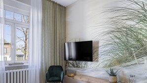A modern living room with a large window, a flat-screen TV mounted on the wall, a green chair, a wooden bench, and a potted plant.