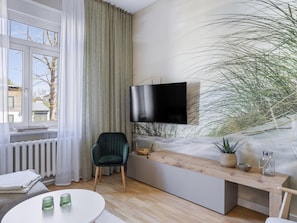 Ein modernes Wohnzimmer mit großem Fenster, einem an der Wand befestigten Flachbildfernseher, einem grünen Sessel, einer Holzbank und einer Topfpflanze.