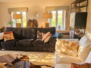 A living room with a dark-colored sofa, a white sofa, and a coffee table. There are two windows with curtains, a fireplace, and a bookshelf with decorative items. - Authentic Charente house in a quiet natural area (La Villedieu)