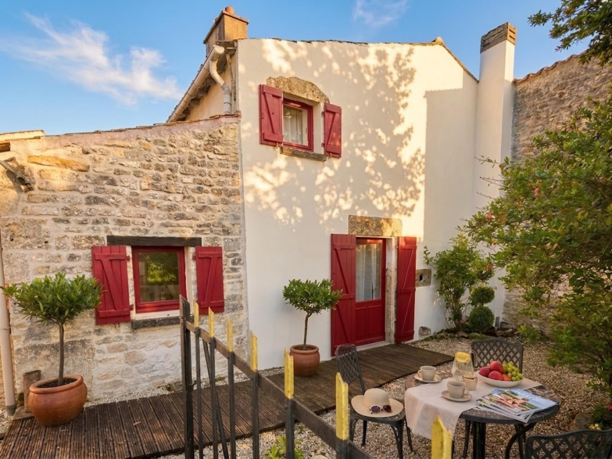 A stone building with red shutters, a small garden with a table set for tea, and a wooden deck.