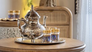 Une table avec une théière en argent, des tasses et un plateau, placée devant un mur décoré de guirlandes lumineuses et agrémentée d’une plante.