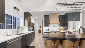 A modern kitchen with dark cabinets, a large island with bar stools, a built-in oven, and a stainless steel refrigerator. - Juniper Presents: NEW Luxe Stay w Hot Tub, Fire Pavilion Nr Lake MI (Berrien County)