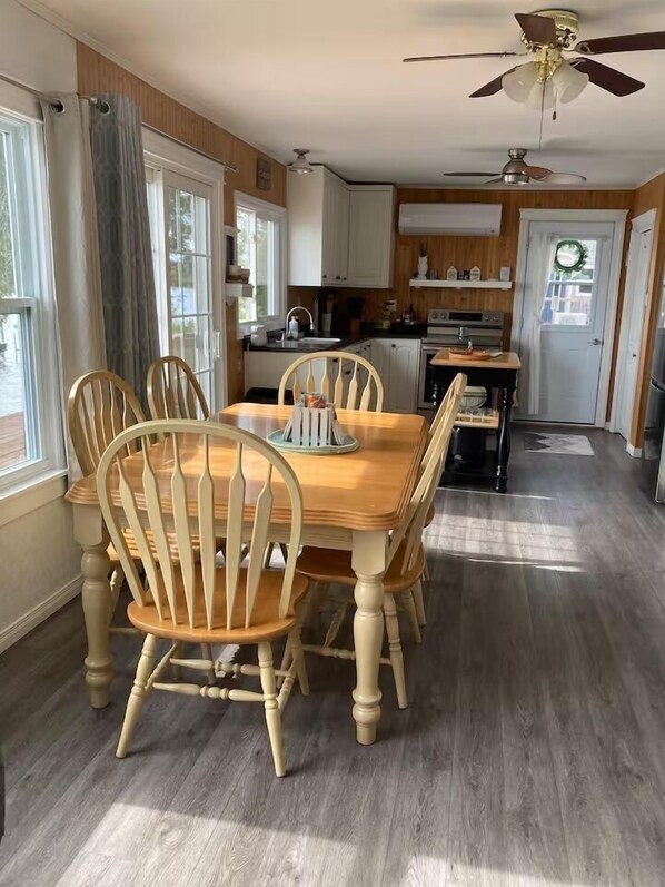 A dining area with a wooden table and chairs, a kitchen in the background, and a ceiling fan.