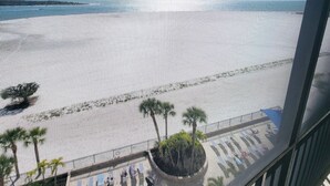 Vista a la playa desde un balcón con barandilla, palmeras y sillas de descanso.