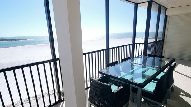 Un balcón con mesa y sillas de vidrio, vista a la playa y piso de baldosas.