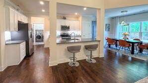 A modern kitchen with a central island, a dining area with a table and chairs, and a view of the outdoors through large windows.