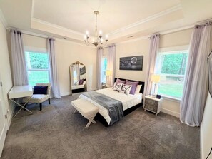 Une chambre à coucher comprenant un lit, des tables de chevet, un bureau, une chaise, un lustre et de grandes fenêtres avec des rideaux.