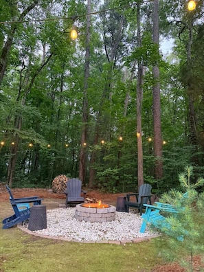 A forested outdoor area with a fire pit, seating, and string lights.