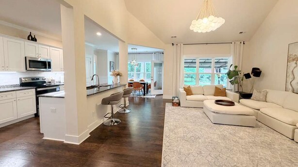 A modern kitchen with white cabinets, a dining area with a table and chairs, and a living room with a white sofa and a large window.