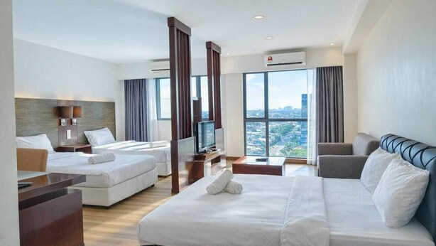 A hotel room with a large bed, two smaller beds, a desk, a TV, and a view of the city.