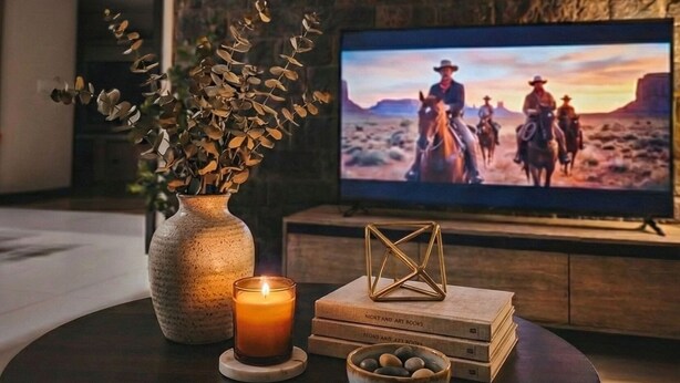 Un salon chaleureux avec un mur en pierre, un téléviseur à écran plat diffusant une scène de western, un vase rempli de fleurs séchées, une bougie allumée et une pile de livres.