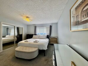 A bedroom with a bed, a mirror, a dresser, a bedside table, and a window with curtains.
