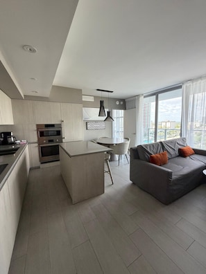 Eine moderne Küche mit einer zentralen Kochinsel, einem Essbereich mit rundem Tisch und einem grauen Sofa mit orangefarbenen Kissen.