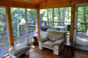 Eine Holzterrasse mit gepolstertem Sitz und einem Tisch, umgeben von großen Fenstern mit Blick auf ein bewaldetes Gebiet.
