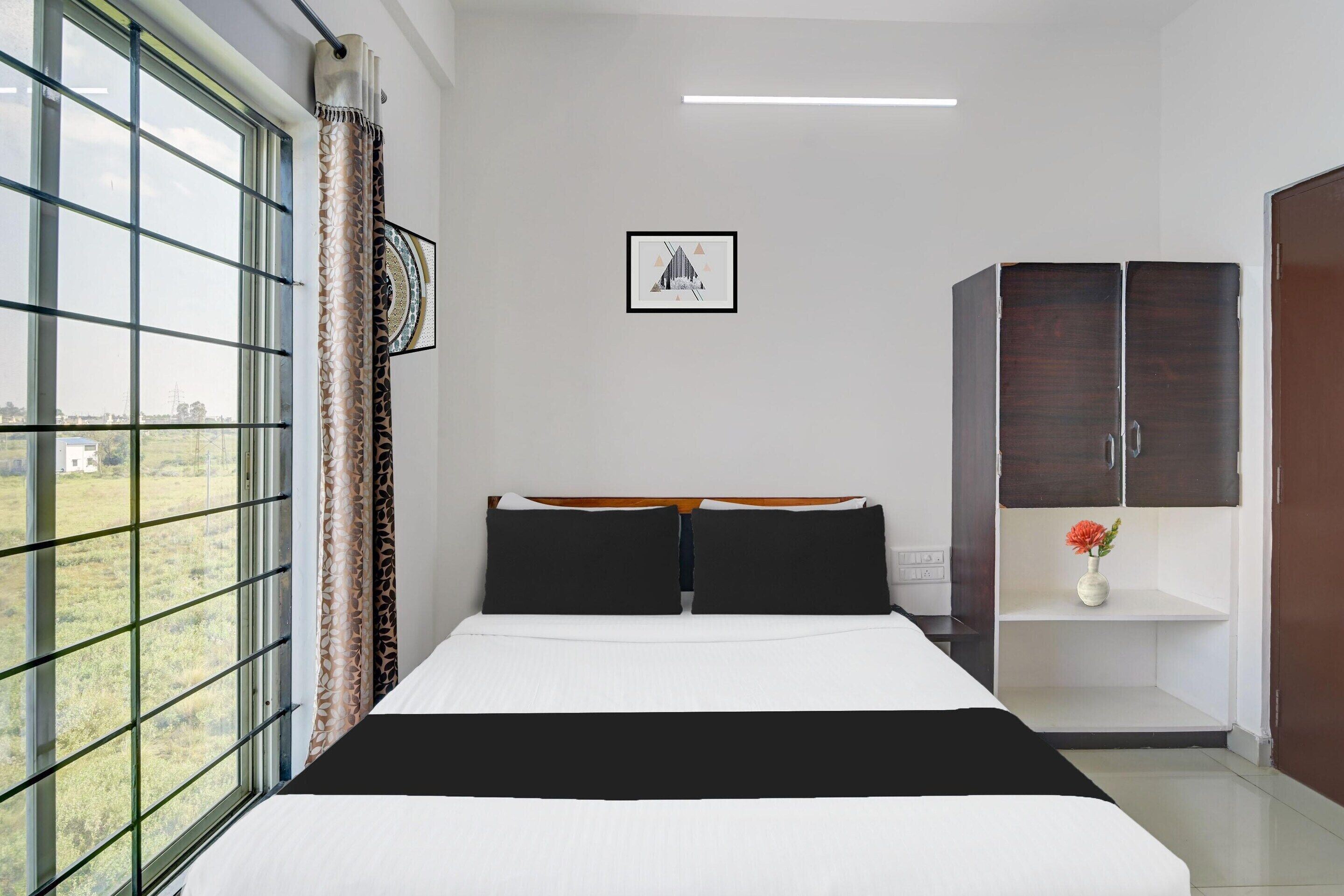 A bedroom with a large window, a bed with black and white bedding, a dark wardrobe, and a small shelf with a vase.