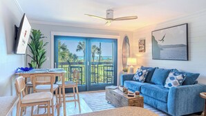 Un salon avec un canapé bleu, des chaises en bois, une table basse, un téléviseur fixé au mur et un balcon offrant une vue sur des palmiers.