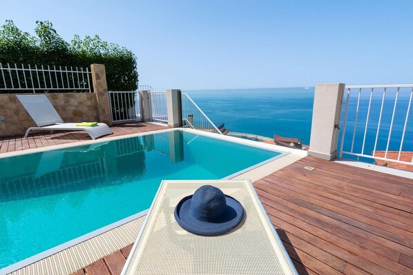 A pool with a view of the sea, a wooden deck, and a lounge chair with a hat.