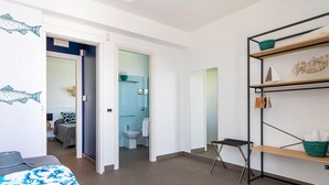 Ein modernes Hotelzimmer mit einem Bett, einem kleinen Schreibtisch, einem Badezimmer inklusive Toilette und Waschbecken sowie einem Regal mit Handtüchern und einem Wäschekorb.