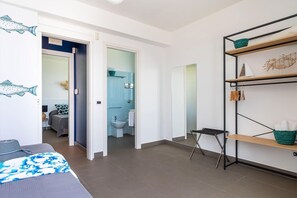 A modern hotel room with a bed, a small desk, a bathroom with a toilet and sink, and a shelf with towels and a basket.
