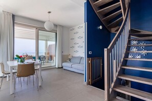 A modern living room with a sofa, dining table, chairs, a staircase, and a view through a glass door.