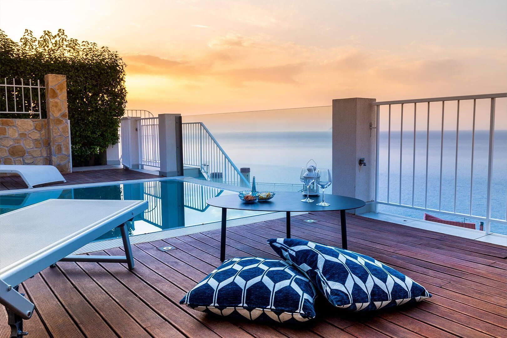 A deck with a pool, a table with glasses and a bowl, and two patterned cushions.