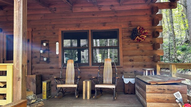 Una cabaña de madera con dos mecedoras, una mesa de madera y una ventana con vista a un bosque.